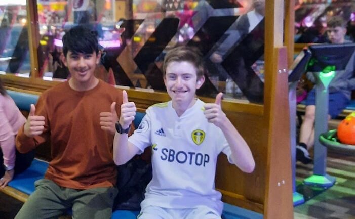 Two boys sit on a bench at a bowling alley looking at the camera smiling with their thumbs up.