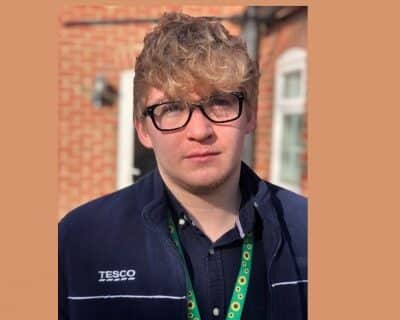 Young white man with short, wavy blonde hair and glasses wearing a Tesco uniform.