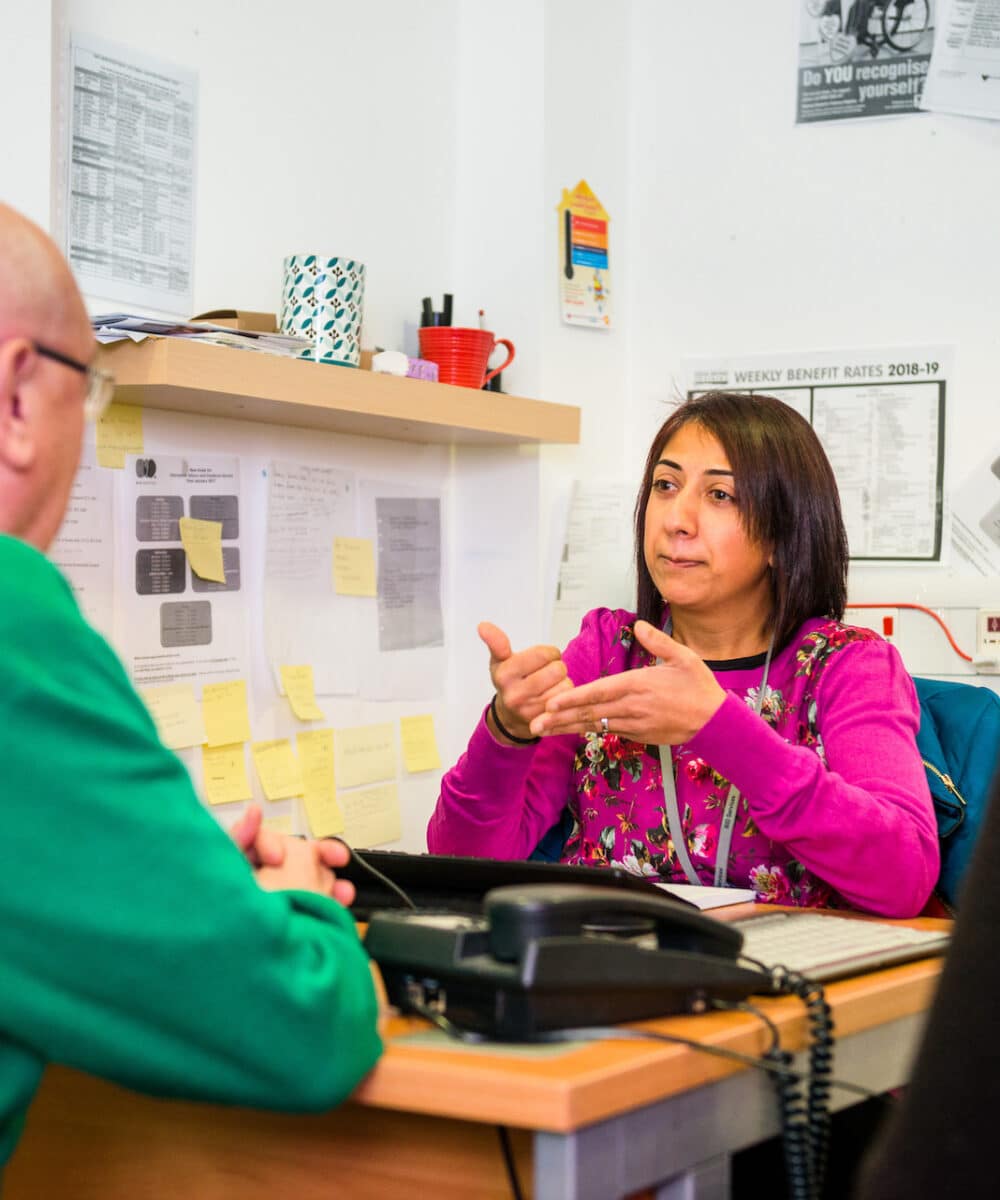 A lady sits at a desk signing to an older man on the other side of the table.