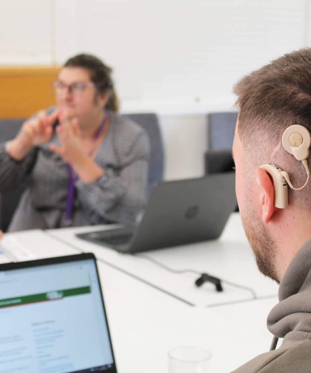 A man with a cochlear implant sits at a table looking at a laptop. Around the table in the background are 3 other people one of which is signing to another.