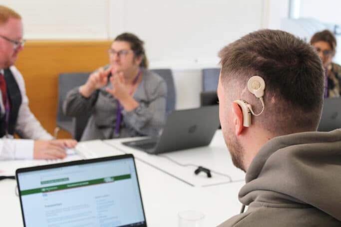 A man with a cochlear implant sits at a table looking at a laptop. Around the table in the background are 3 other people one of which is signing to another.