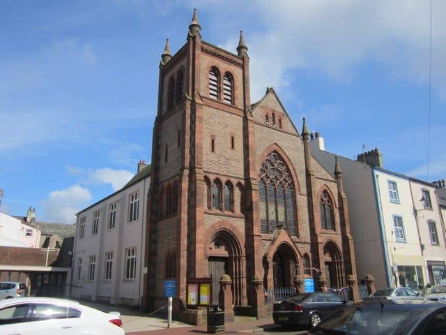 An old brown church, United Reformed Church Whitehaven, sits in the centre of the photo with a white building coming off the back of the church front and other shops sitting to the right.