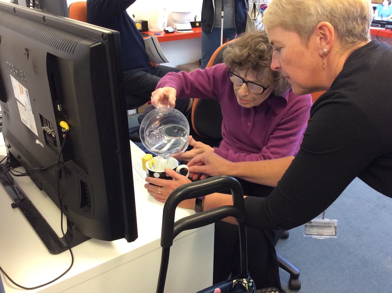An elderly woman is being supported in using a liquid level indicator. She is pouring a jug of plain water into a mug.
