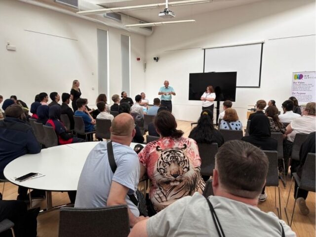 A room full of people, all facing forward towards the front of the room where a man is giving a speech in BSL.