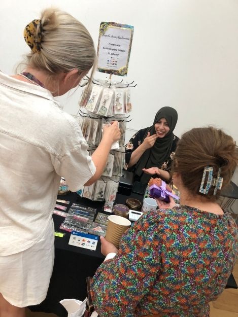 A woman is having a chat with a customer at her handmade gifts stall. Another potential customer is looking through keychains.