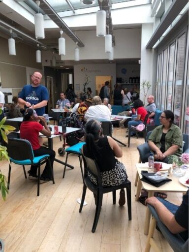A cafe area with over 15 people seated around various tables, eating food.