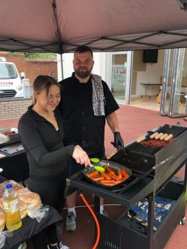 A chef man and woman are stood under a gazebo, frying sausages on griddles and pans.