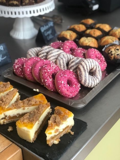 A tray of desserts laid out on silver and black trays. On the left are slices of a fruity cheesecake. In the centre are 3 alternating rows of pink and white chocolate & dark chocolate glazed donuts. To the right are blueberry and plain muffins.