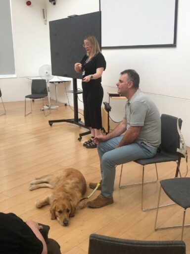 Alex is seated at the front of a room with his guide dog, Ned laying by his feet. He is talking to people off-camera. Beside him is a female BSL interpreter.