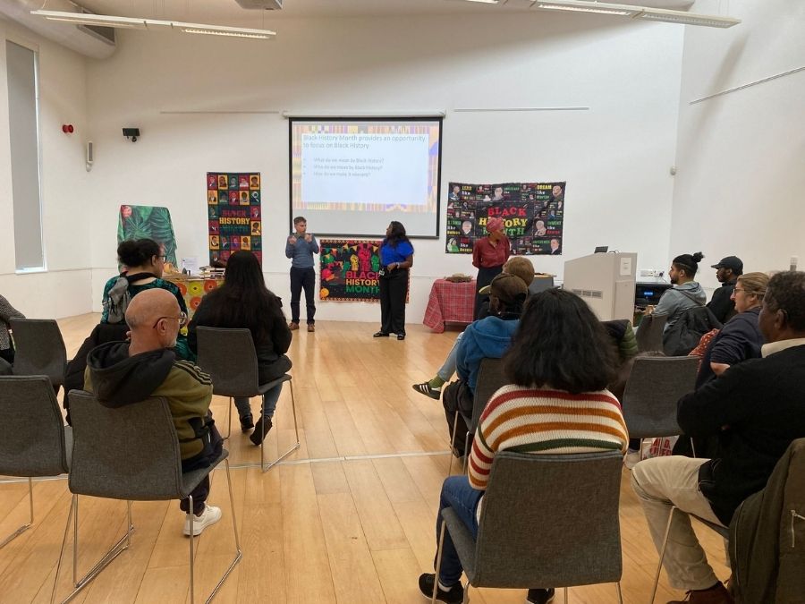 Rows of people seated, facing towards a large screen and 2 presenters at the front. They're celebrating Black History Month.