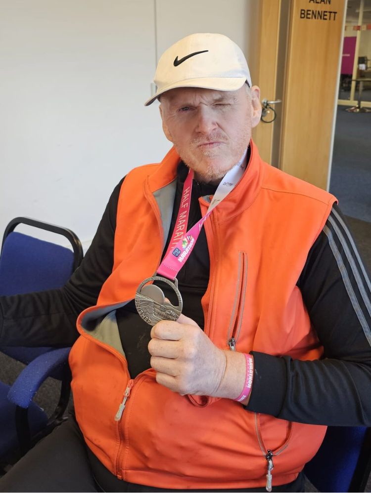 Tim, a white man is seated, wearing a fluorescent orange jacket and holding up his half marathon medal.