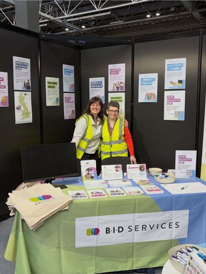 Georgia and Lisa are stood together behind their table in yellow hi-visibility jackets and smiling. The stand is set up with many branded items including a table cloth, leaflets and tote bags. Behind them on the wall are lots of posters.