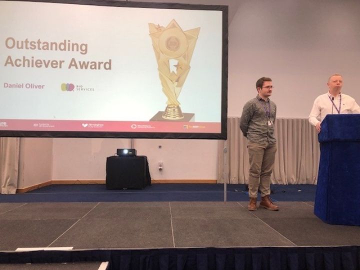 Daniel and Jock are stood by a podium on stage. Behind them is a large screen with the words 'outstanding achiever award' for Daniel.