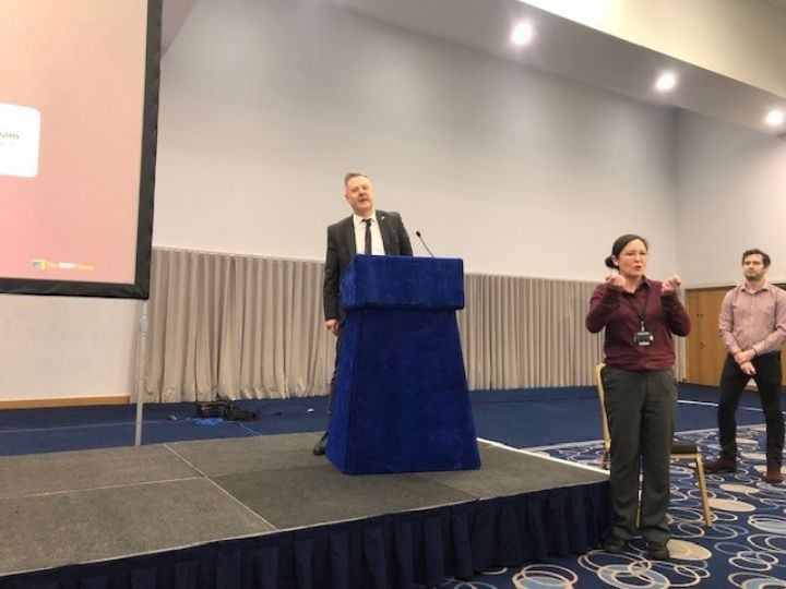 A man is stood behind a podium, giving a speech. In front is a female bsl interpreter.