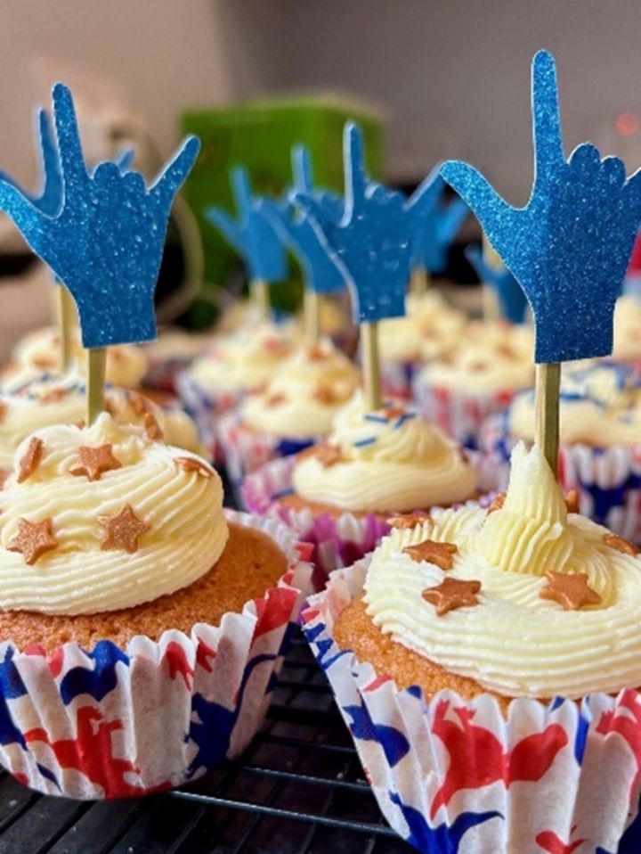 A batch of cupcakes in white, red and blue cases. They're topped with white buttercream swirls, gold star sprinkles and glitter card cupcake toppers which spell out 'i love you' in american sign language.