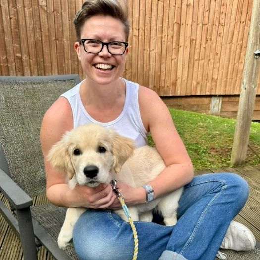 Sarah, a white woman with short hair and glasses is sat cross legged with a Labrador retriever puppy on her lap.