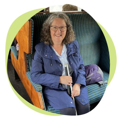 A white woman with shoulder length curly grey hair. She is seated on a train and holding a white cane.