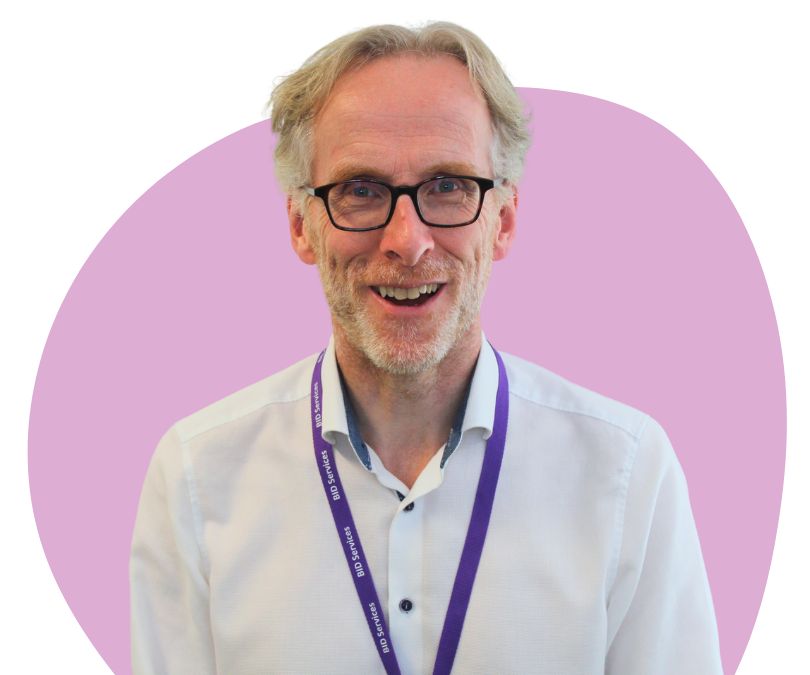 White mane with short light grey hair with black framed glasses against a purple background.