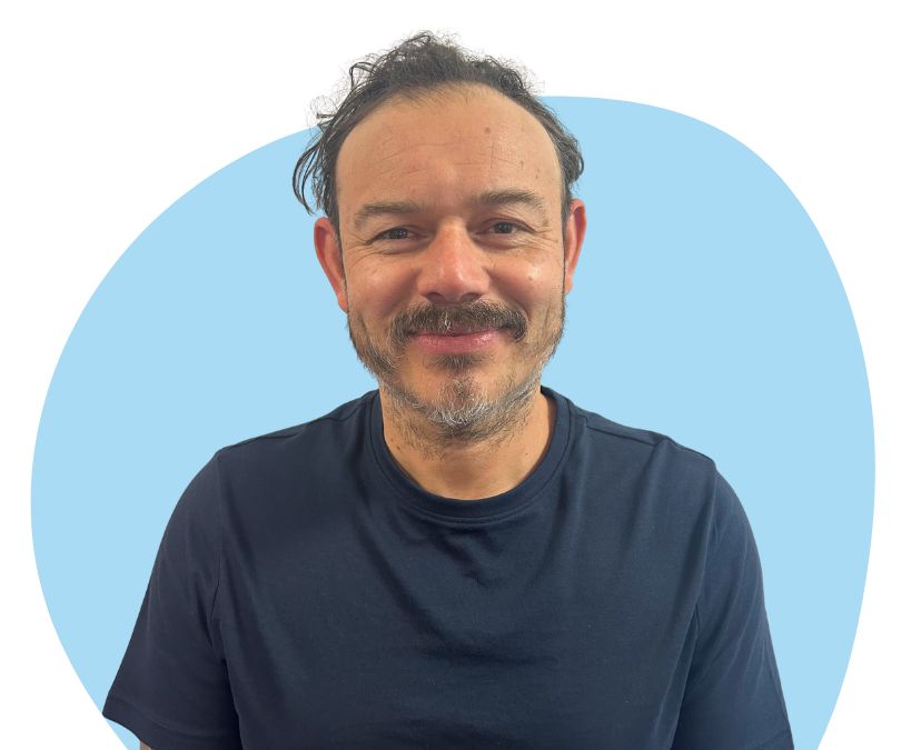 A white man with short black hair and facial hair against a light blue background.