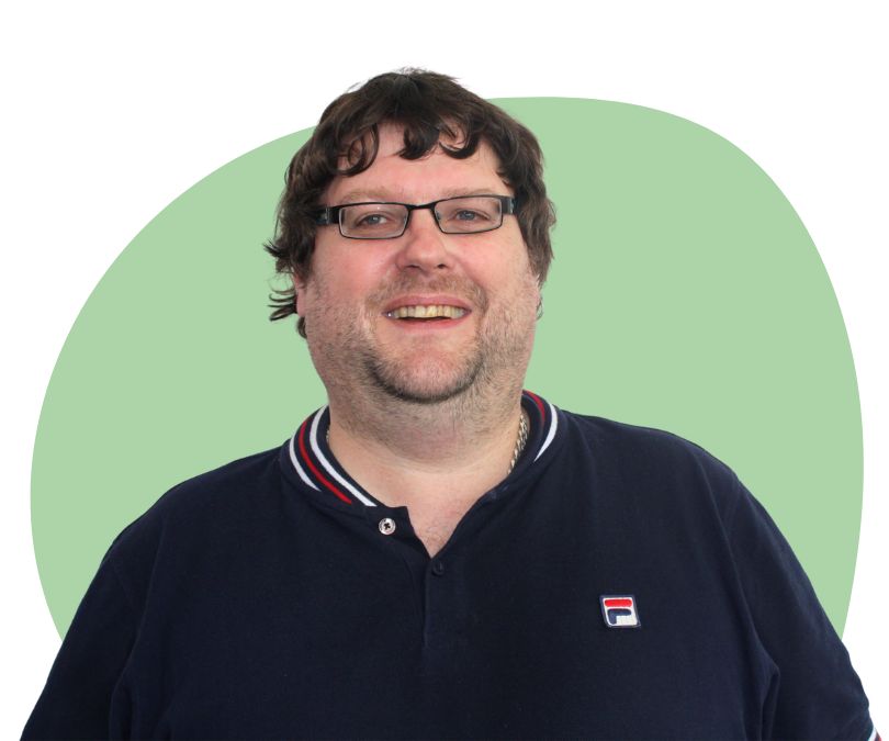 White man with short brown hair and stubble and glasses against a green background,