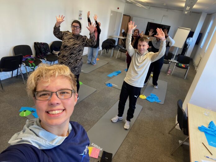 A selfie of a blonde woman. In the background are 5 men and women standing on exercise mats with their hands up in the air and smiling towards the camera.