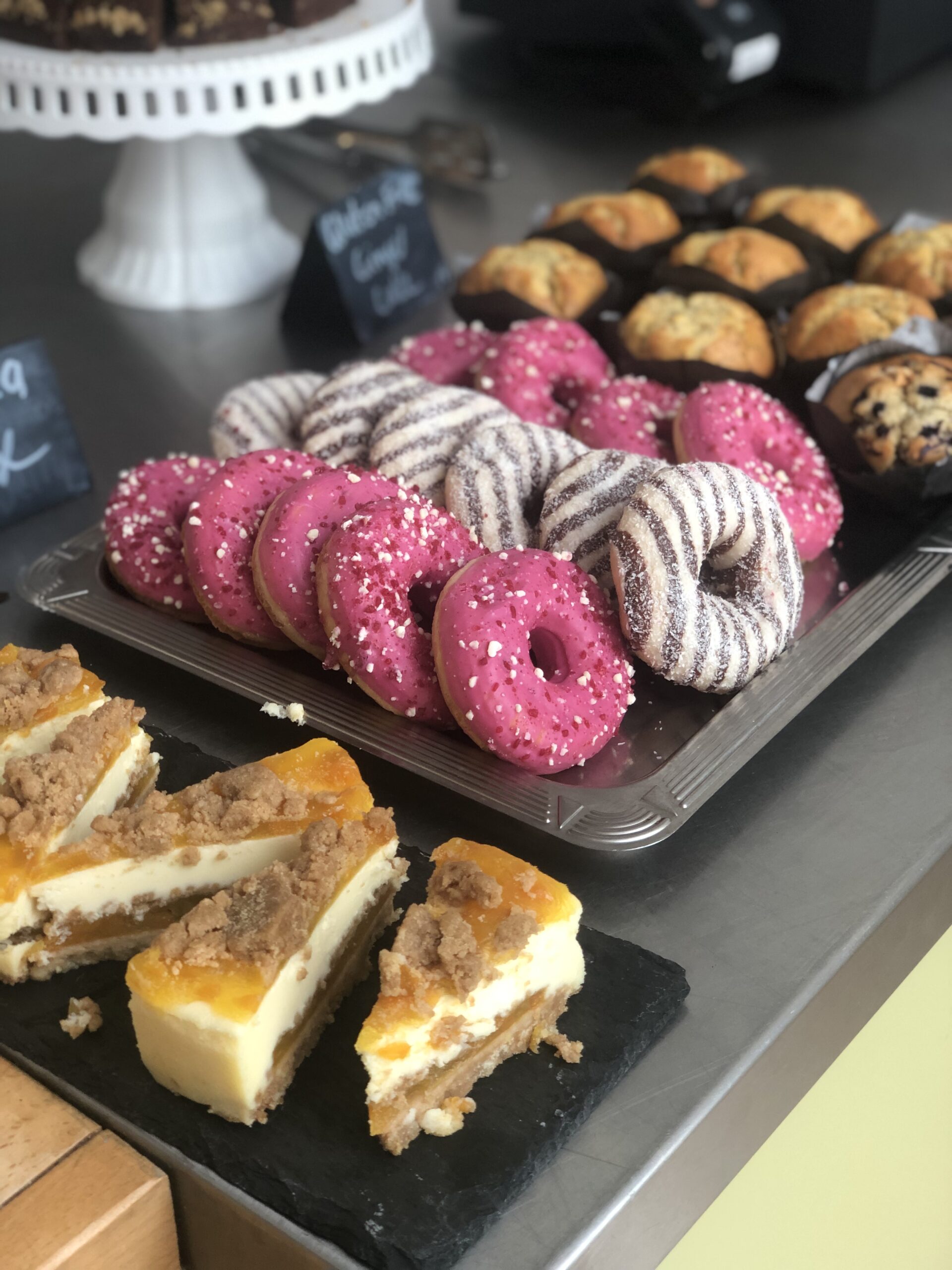 A tray of desserts laid out on silver and black trays. On the left are slices of a fruity cheesecake. In the centre are 3 alternating rows of pink and white chocolate & dark chocolate glazed donuts. To the right are blueberry and plain muffins.
