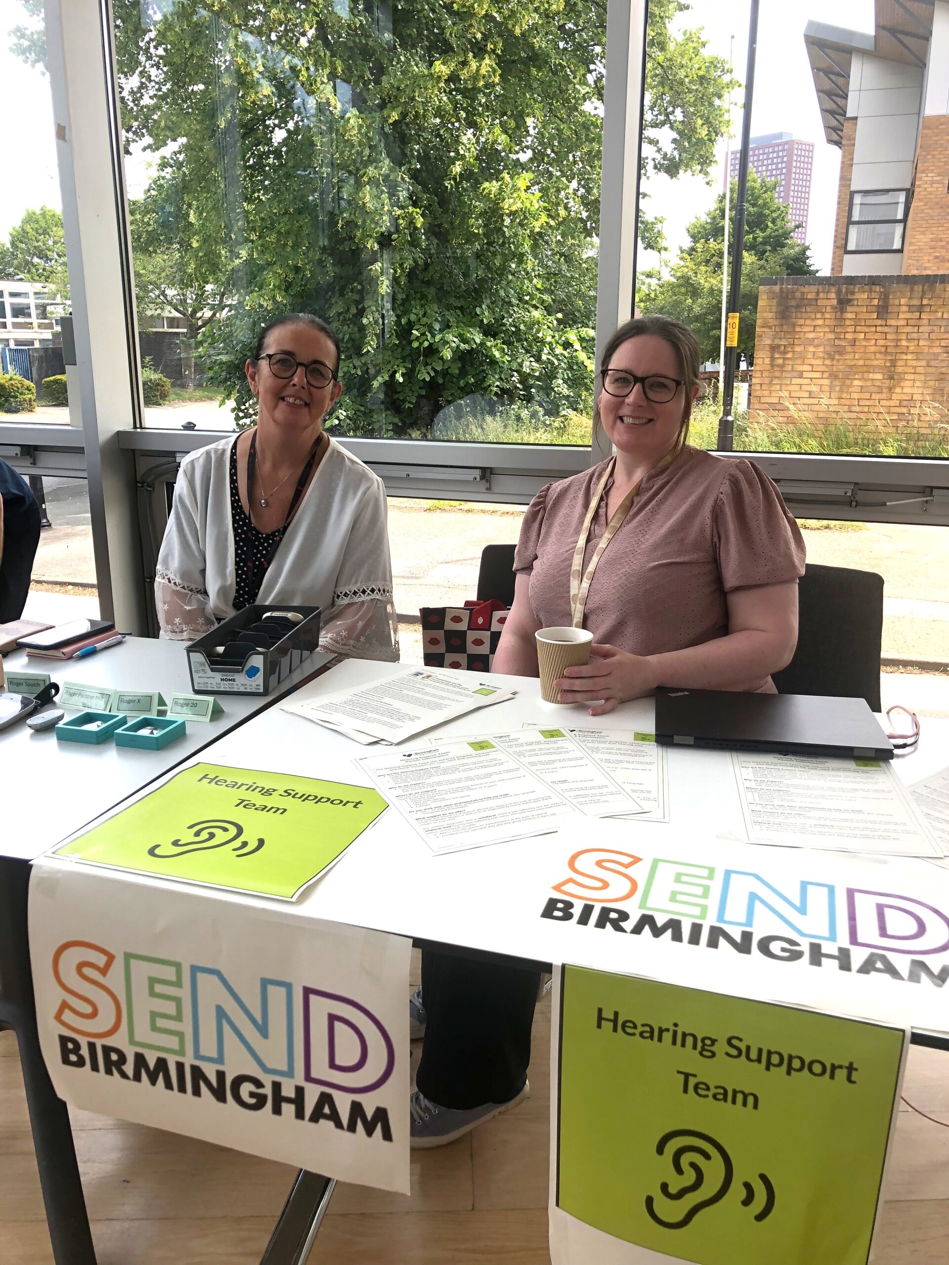 2 white females are seated behind their stall for SEND Birmingham. They're both smiling towards the camera.