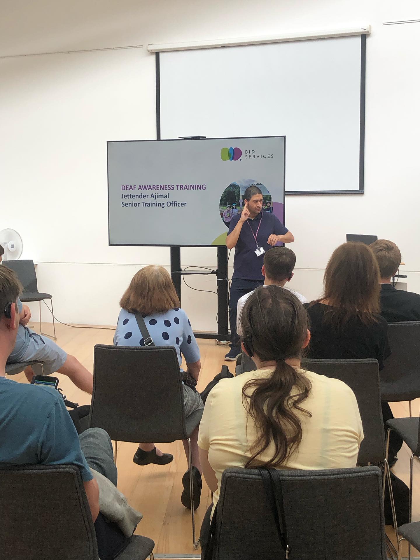 An Asian man is stood by a large TV screen, delivering a presentation for deaf awareness training.