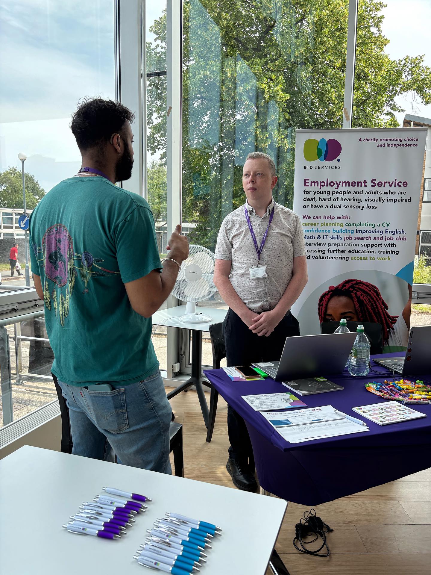 2 men are stood and in conversation in British sign Language, stood by an employment service stall.