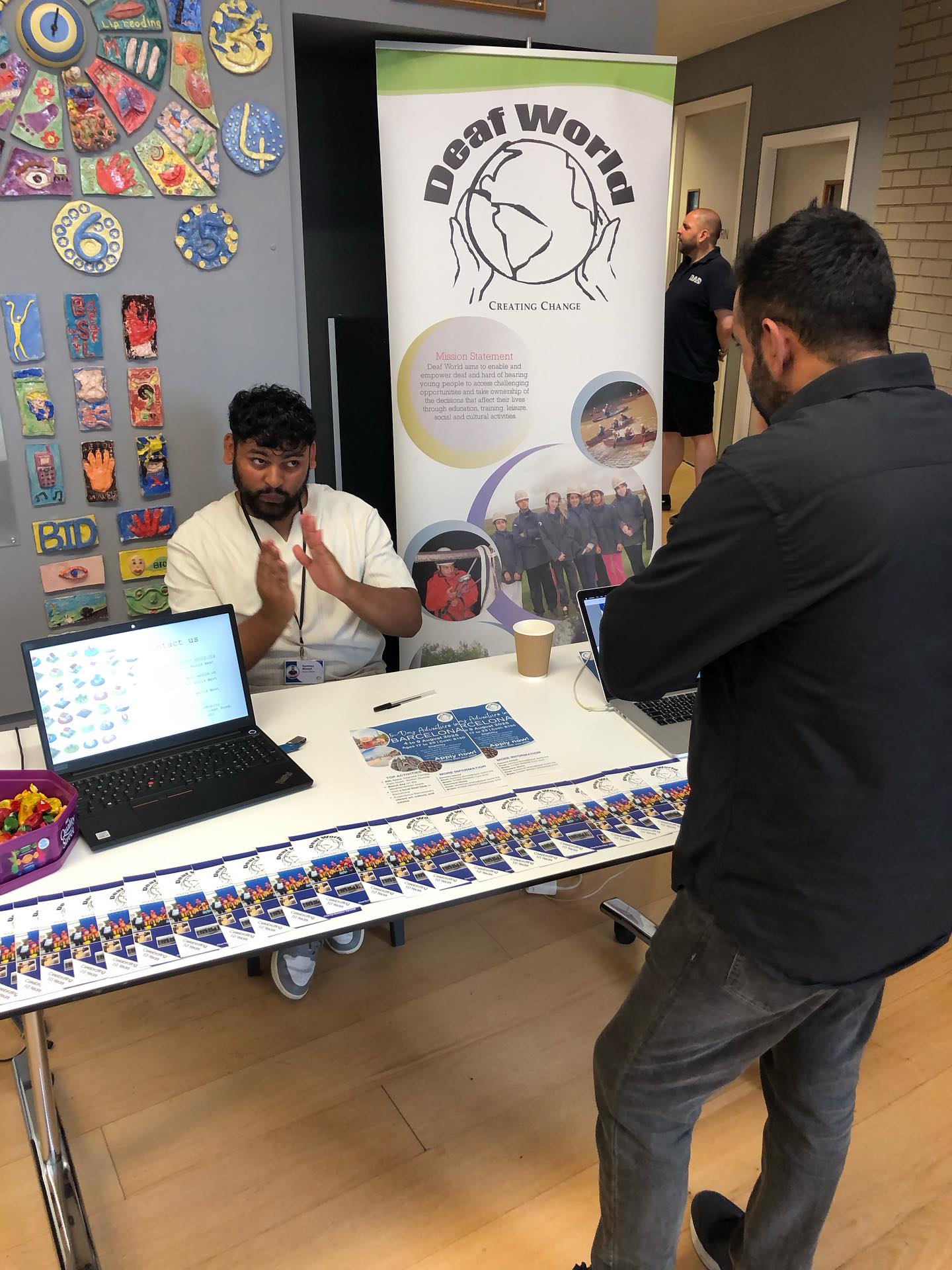 An Asian man representing Deaf World is seated by his stall. He is using British Sign Language to communicate with a man stood opposite him.