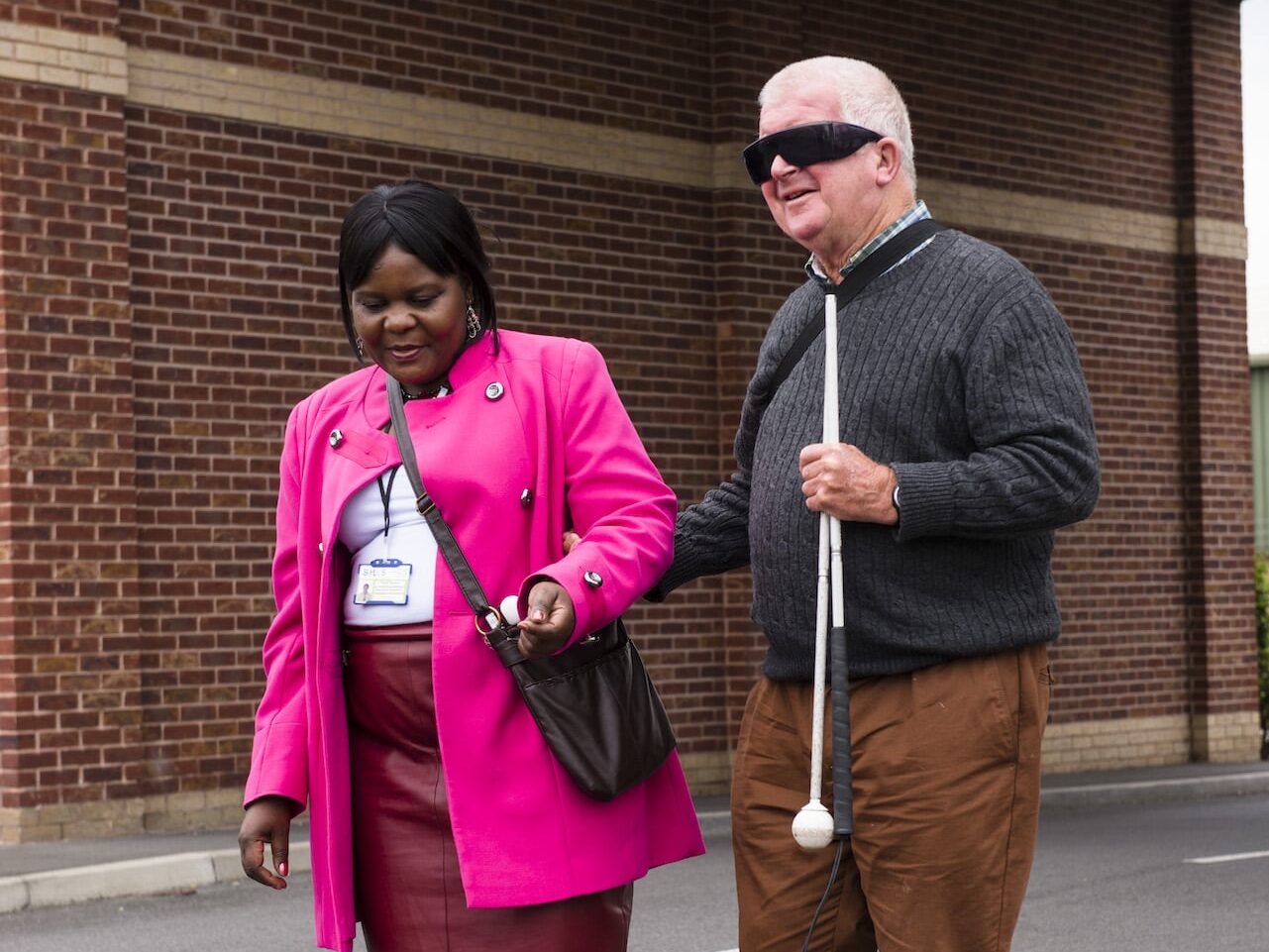 A man with dark glasses and a folded up cane in his hand holds onto the elbow of a lady as they start crossing a road.