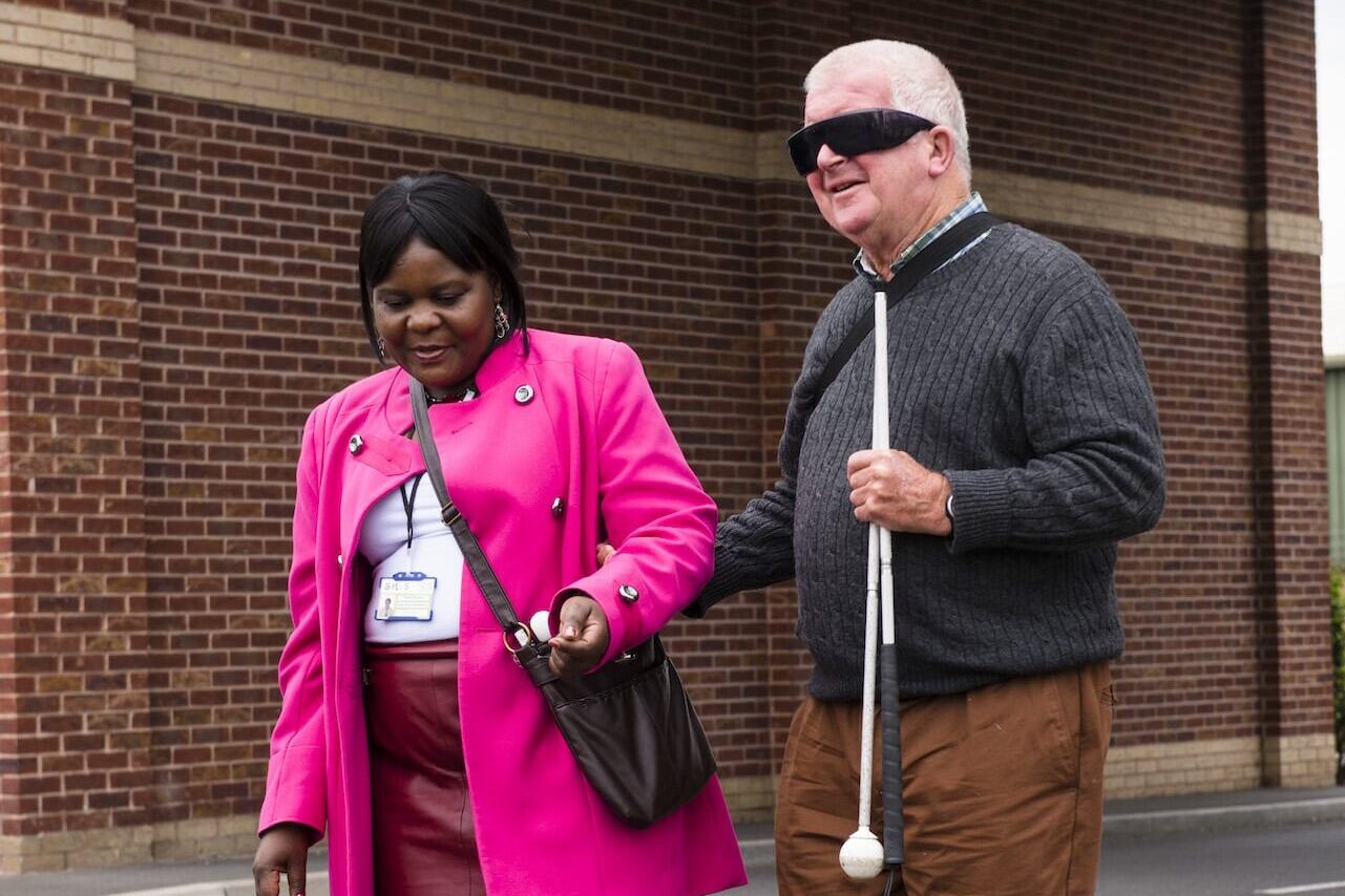 A man with dark glasses and a folded up cane in his hand holds onto the elbow of a lady as they start crossing a road.