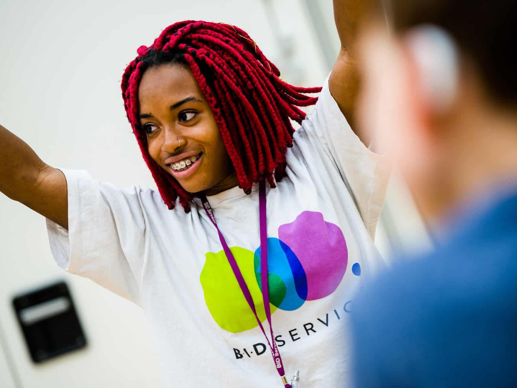 A lady with a BID Services t-shirt on smiles looking away from the camera with her hands in the air.