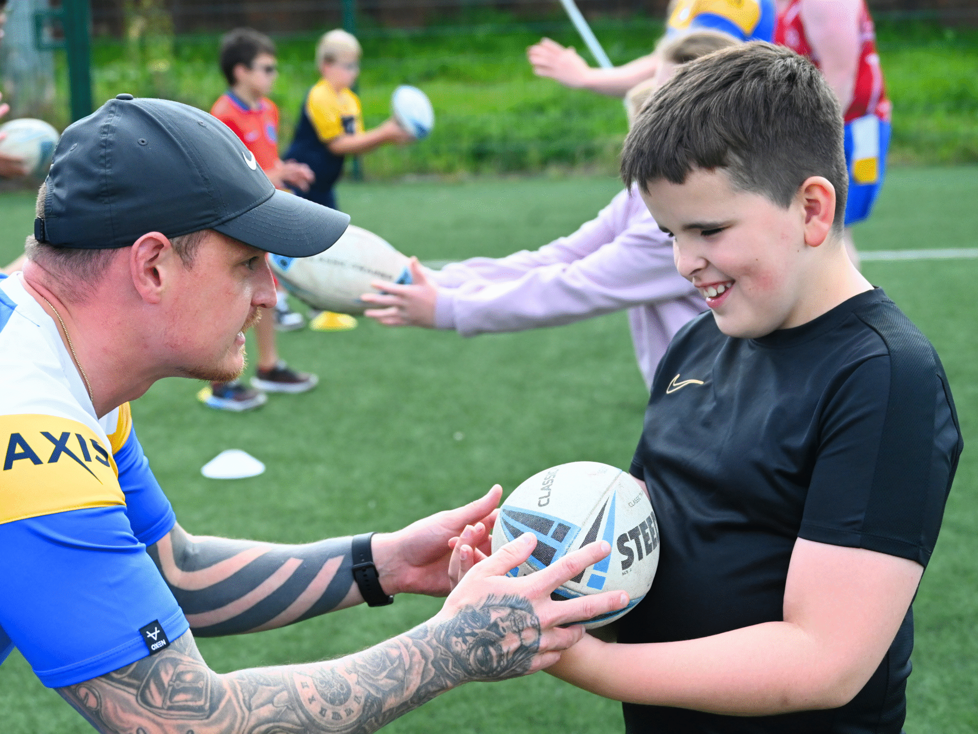 On a rugby practice pitch, a young boy is smiling while being handed a rugby ball by an older man.