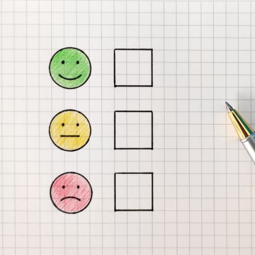 Three hand‑drawn faces arranged vertically on grid paper: a green smiling face at the top, a yellow neutral face in the middle, and a red sad face at the bottom. To the right of each face is an empty checkbox. A pen rests on the right side of the page.