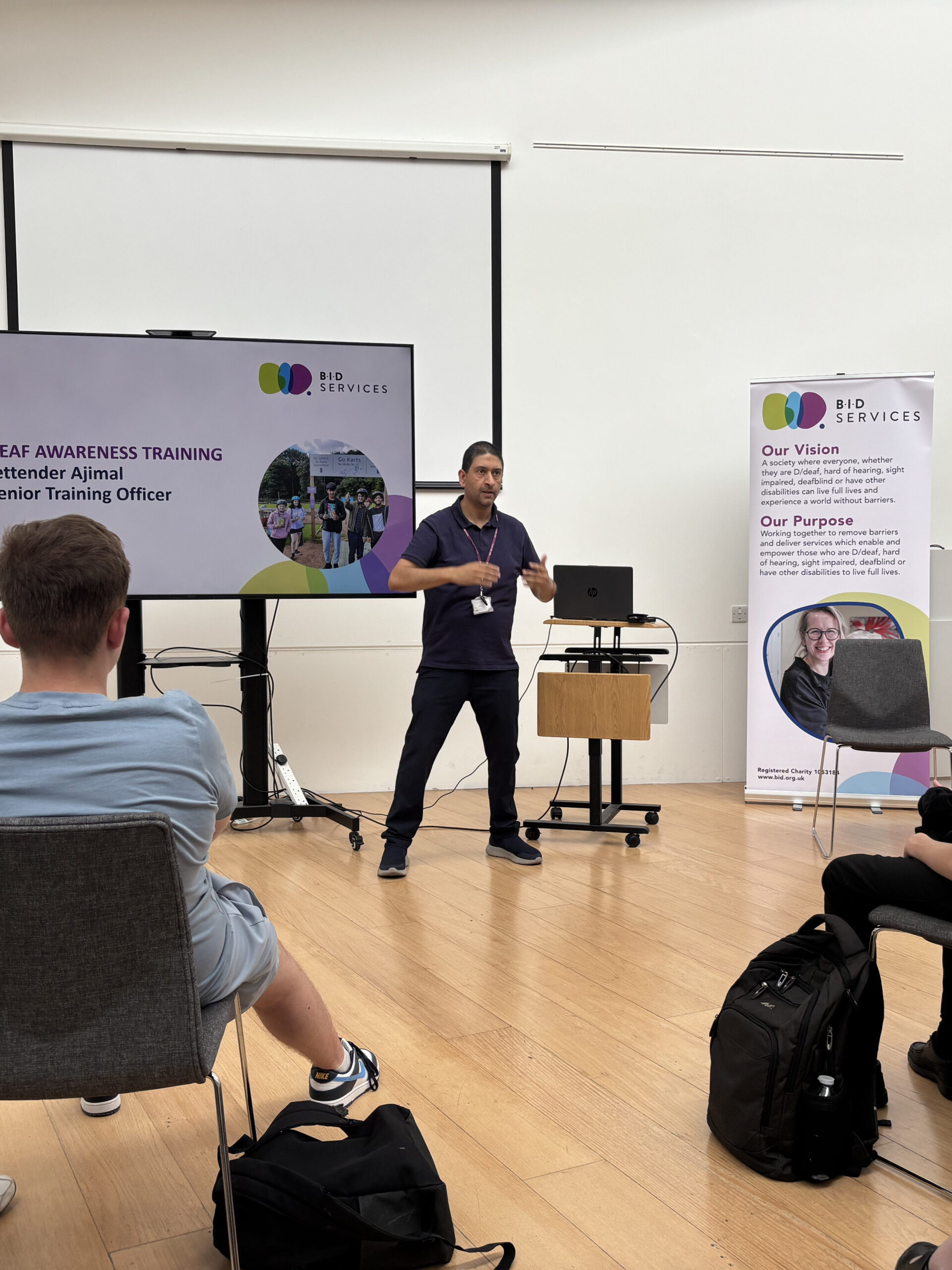 An Asain man stood by a large screen, giving a presentation on Deaf Awareness.