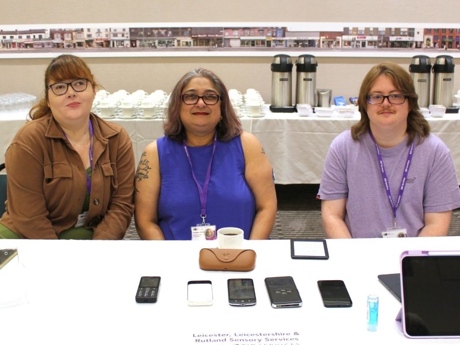 2 female and a male member of staff are seated beside each other behind a table which is filled with phones and tablets. They are all looking towards the camera and smiling.