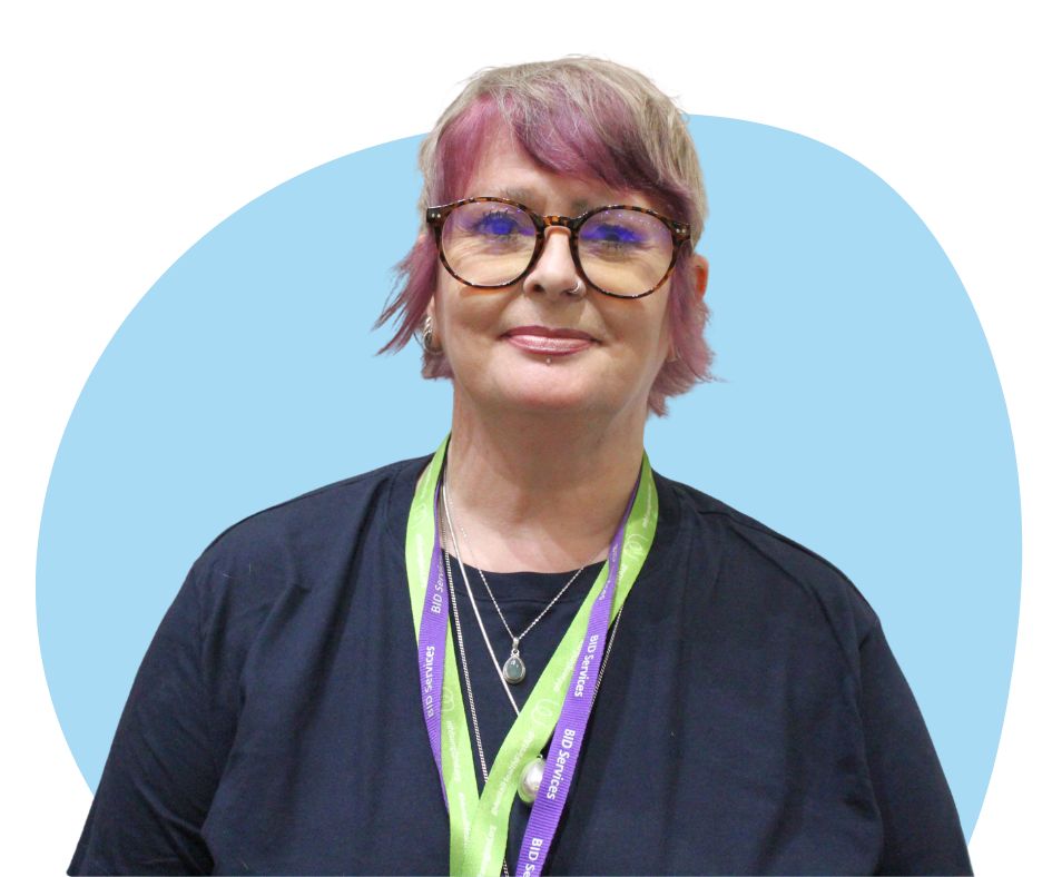 White woman with short grey and purple hair with large round glasses against a light blue background.